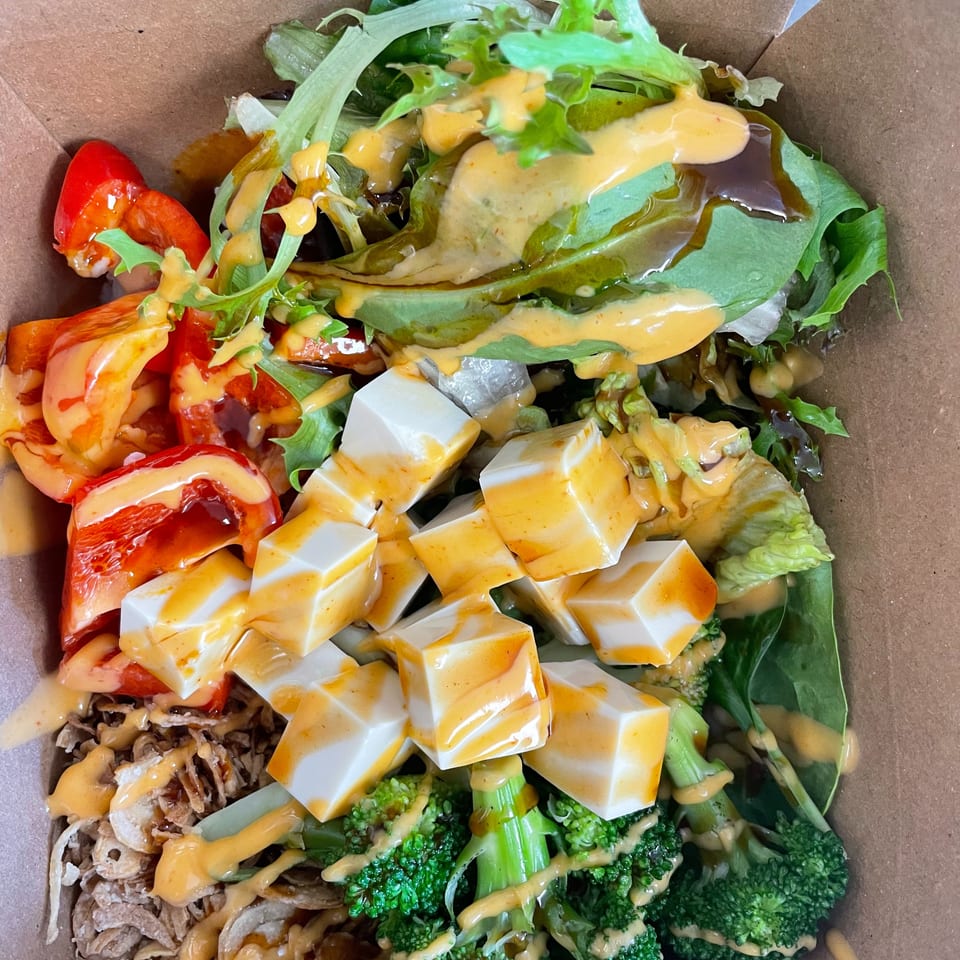 Colorful vegan bowl with tofu, red peppers, broccoli, and greens, drizzled with two sauces in a takeout box.
