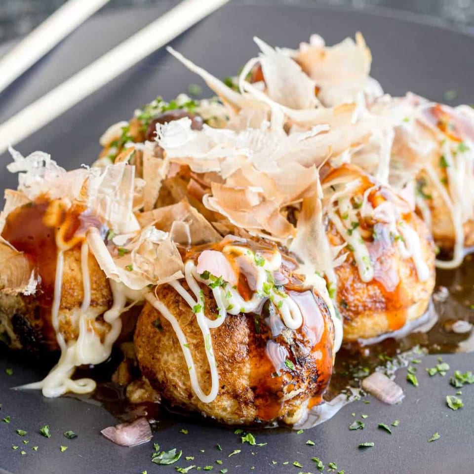 Takoyaki with savory sauce, mayonnaise, bonito flakes, and green herbs on a dark plate.