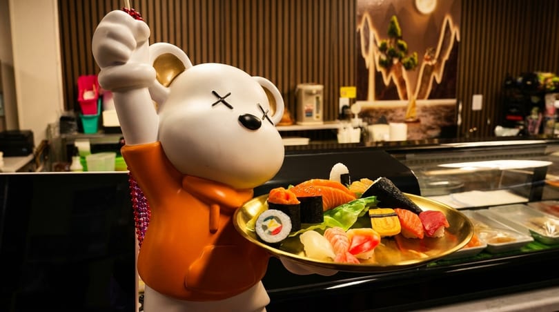 A white bear statue in an orange hoodie holds a gold tray with various sushi pieces.