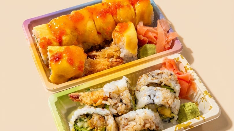 Two takeout containers of sushi rolls, one with orange sauce and the other with sesame seeds.